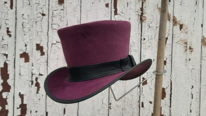 May include: A burgundy top hat with a black leather band. The hat is displayed on a wooden stand against a white wooden background.