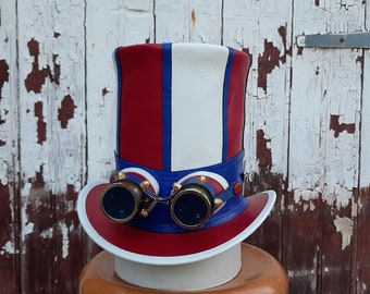 May include: A red, white, and blue top hat with a pair of steampunk goggles attached to the front. The goggles have gold accents and blue lenses.
