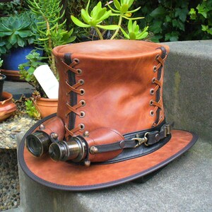 May include: A brown leather top hat with a black band and brass accents. The hat has a pair of brass binoculars attached to the front.