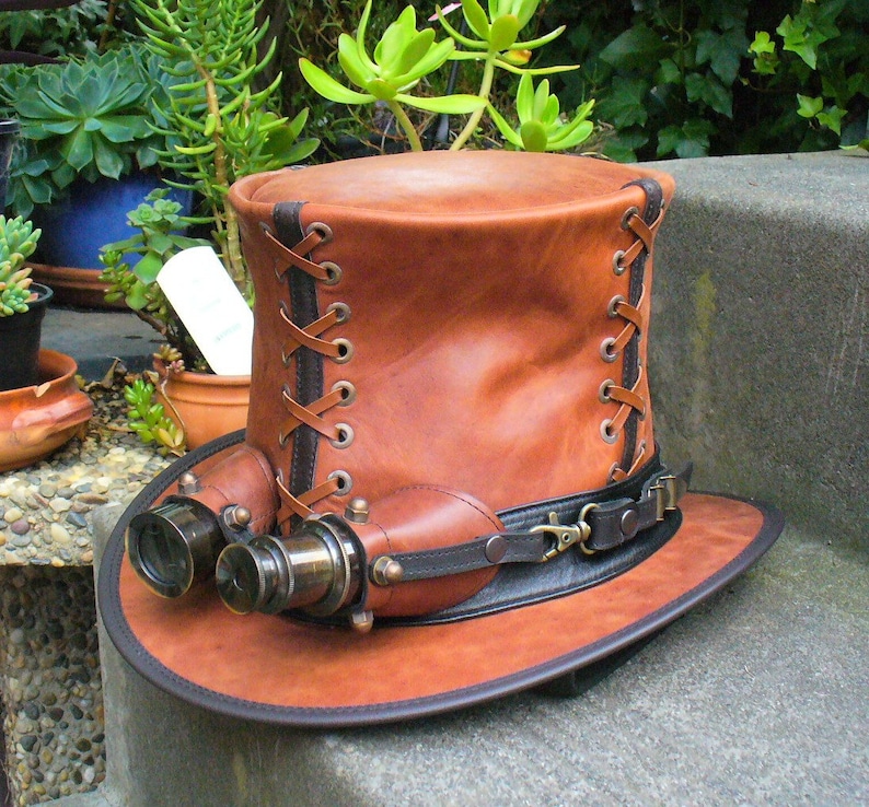 May include: A brown leather top hat with a black band and brass accents. The hat has a pair of brass binoculars attached to the front.