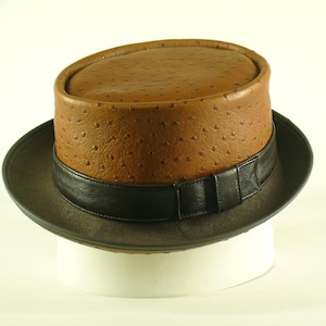 Puede incluir: Un sombrero fedora de cuero marrón con una banda negra. El sombrero tiene un patrón texturizado, similar al del avestruz.