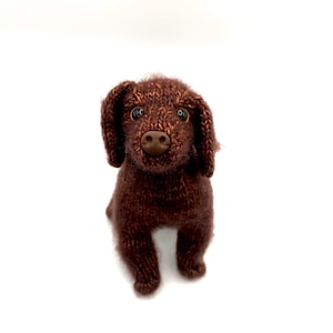May include: A brown knitted dog toy with a button nose and floppy ears. The dog is sitting upright, with its front paws visible. The toy is made of a textured yarn and has small, dark eyes.