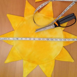 Op de afbeelding: Een geel, zonvormig stuk stof met een wit tie-dye patroon. De stof wordt gemeten met een meetlint en geknipt met een schaar.