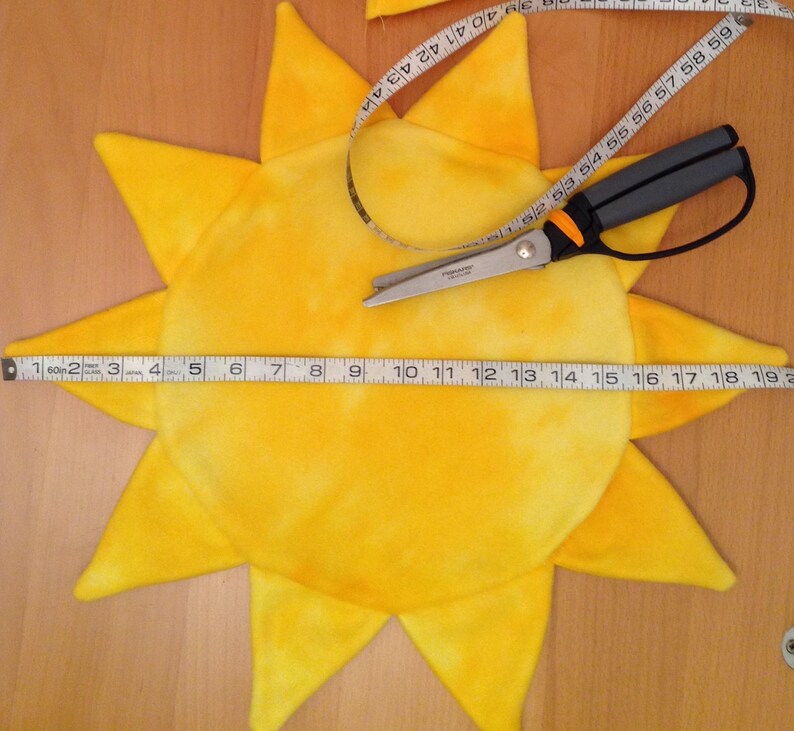 Op de afbeelding: Een geel, zonvormig stuk stof met een wit tie-dye patroon. De stof wordt gemeten met een meetlint en geknipt met een schaar.