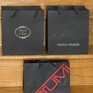 May include: Three black shopping bags with black rope handles. Two bags have the text "VENUS ET FLEUR" in gold. One bag has a diamond pattern. The third bag has the text "TUMI" in red.
