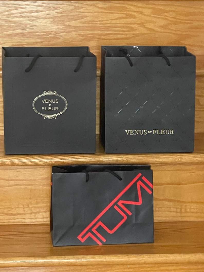 May include: Three black shopping bags with black rope handles. Two bags have the text "VENUS ET FLEUR" in gold. One bag has a diamond pattern. The third bag has the text "TUMI" in red.