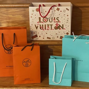 May include: An assortment of luxury shopping bags in various sizes and colors. The bags include orange Hermes bags, a white Louis Vuitton bag with red handles, and Tiffany & Co. bags in a light blue color.