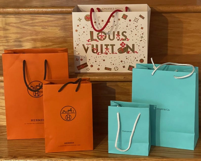 May include: An assortment of luxury shopping bags in various sizes and colors. The bags include orange Hermes bags, a white Louis Vuitton bag with red handles, and Tiffany & Co. bags in a light blue color.