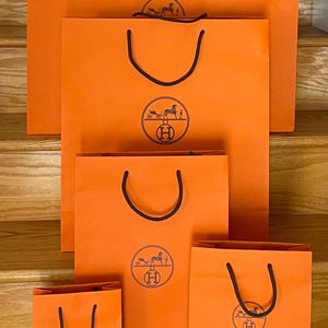 May include: A collection of orange shopping bags of varying sizes, each with black handles and a circular logo featuring a horse-drawn carriage. The bags are arranged on wooden stairs, with the brand name "HERMES" printed on some.