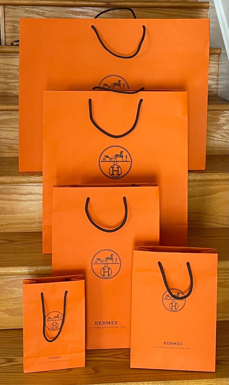 May include: A collection of orange shopping bags of varying sizes, each with black handles and a circular logo featuring a horse-drawn carriage. The bags are arranged on wooden stairs, with the brand name "HERMES" printed on some.