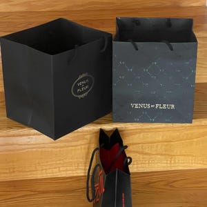 May include: Three black gift bags with handles are displayed on wooden stairs. Two bags are open, revealing their interiors. The largest bag has the text "VENUS ET FLEUR" in gold. The smallest bag has red interior and text.
