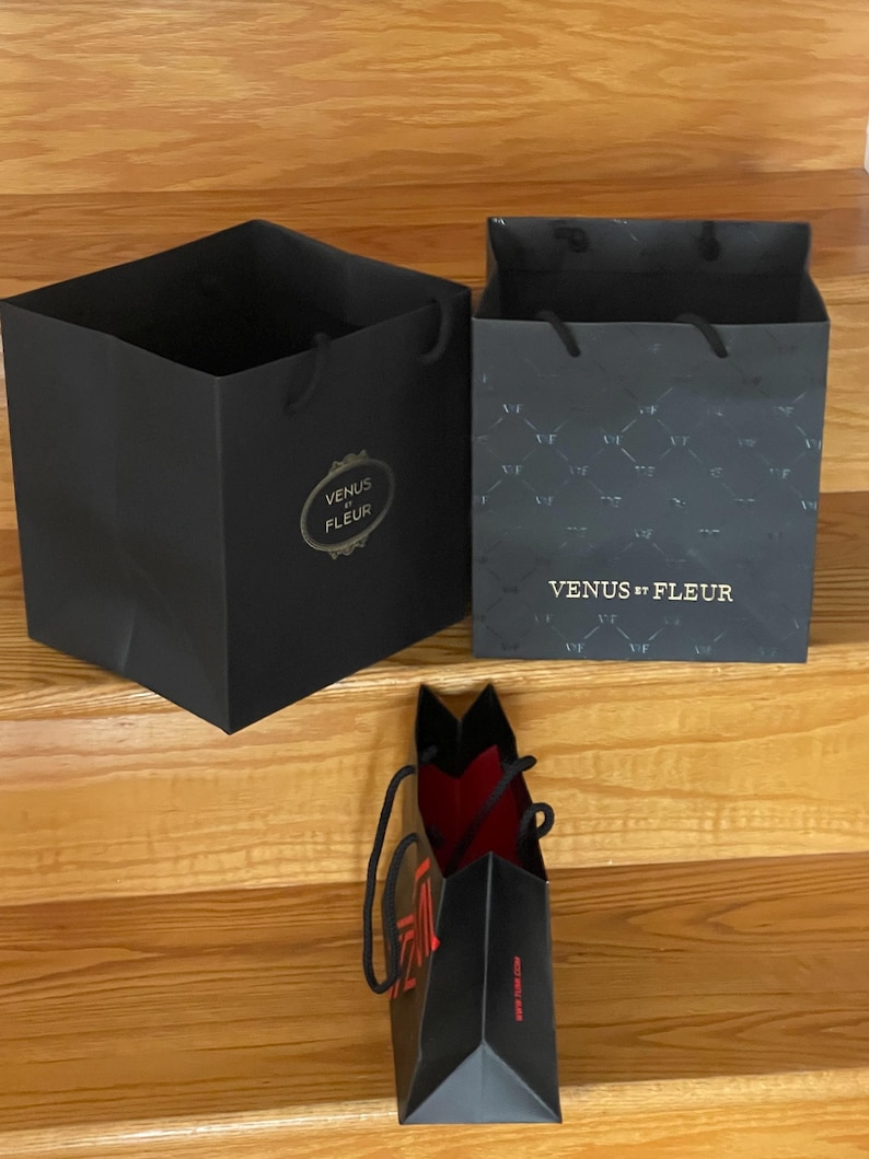 May include: Three black gift bags with handles are displayed on wooden stairs. Two bags are open, revealing their interiors. The largest bag has the text "VENUS ET FLEUR" in gold. The smallest bag has red interior and text.