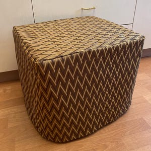 May include: A square ottoman with a brown and gold zigzag pattern. The fabric-covered footstool has a padded top and rounded corners. The design features a repeating chevron pattern in shades of brown and gold.