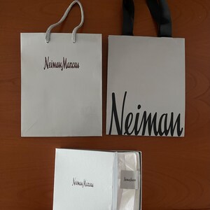 May include: Two white shopping bags and a white box with the "Neiman Marcus" logo. One bag has a silver cord handle, the other has a black handle. The box contains tissue paper and a small tag.
