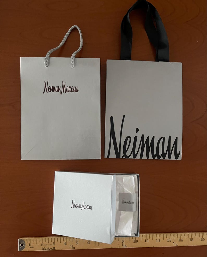 May include: Two white shopping bags and a white box with the "Neiman Marcus" logo. One bag has a silver cord handle, the other has a black handle. The box contains tissue paper and a small tag.