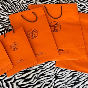 May include: A collection of orange shopping bags with brown handles. Each bag features a circular logo with a horse-drawn carriage and the word "HERMES". The bags are arranged on a black and white zebra-striped surface.