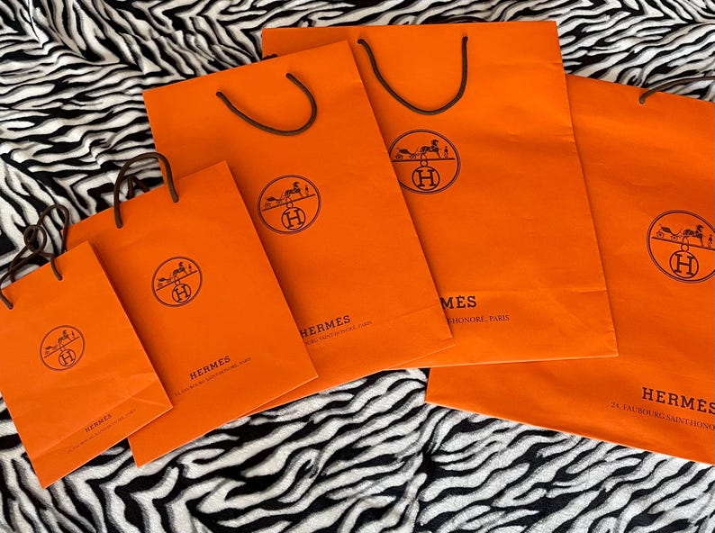 May include: A collection of orange shopping bags with brown handles. Each bag features a circular logo with a horse-drawn carriage and the word "HERMES". The bags are arranged on a black and white zebra-striped surface.