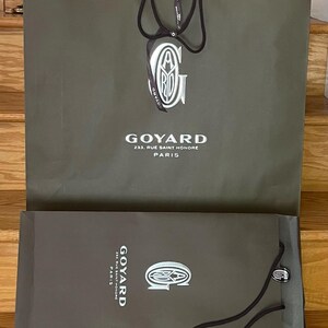 May include: Two olive-green Goyard shopping bags. The larger bag has a white logo and the text "GOYARD 233, RUE SAINT HONORE PARIS". The smaller bag has the same logo and text. Both bags have dark brown handles.