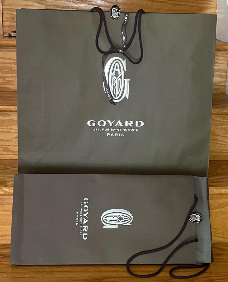 May include: Two olive-green Goyard shopping bags. The larger bag has a white logo and the text "GOYARD 233, RUE SAINT HONORE PARIS". The smaller bag has the same logo and text. Both bags have dark brown handles.