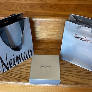 May include: Two gray shopping bags and a box with the name "Neiman Marcus" printed on them. The bags have black handles and the box is light beige. The bags and box are on a wooden surface.