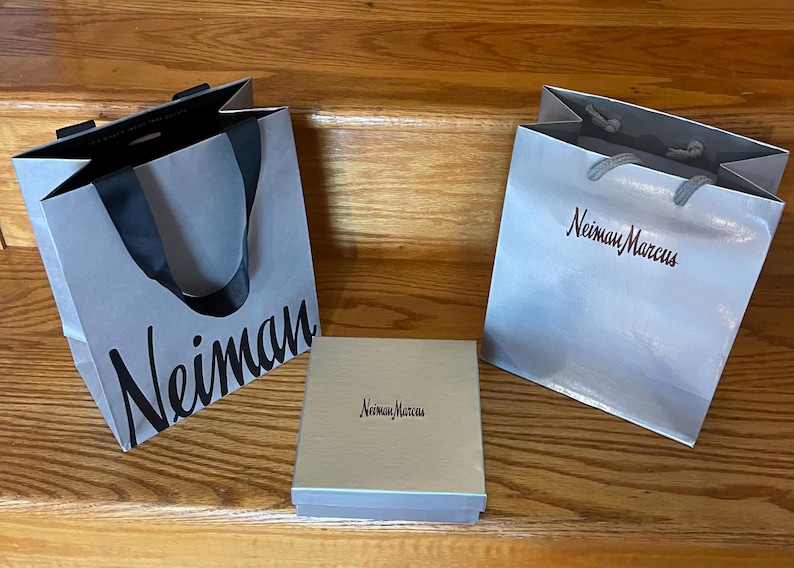 May include: Two gray shopping bags and a box with the name "Neiman Marcus" printed on them. The bags have black handles and the box is light beige. The bags and box are on a wooden surface.