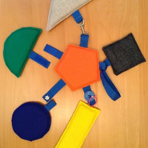 May include: A set of felt shapes connected by blue ribbon and a silver chain. The shapes include a triangle, a semi-circle, a hexagon, a circle, a rectangle, and a square. The shapes are in various colors, including green, orange, blue, yellow, gray, and white.