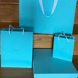 May include: A collection of Tiffany & Co. gift boxes and bags in a signature robin's egg blue. The set includes various sizes of shopping bags with white handles and a rectangular box. The Tiffany & Co. logo is visible on each item.