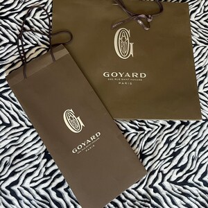 May include: Two brown paper shopping bags with the Goyard logo and address printed in white. The bags have brown handles and are on a black and white zebra-striped background. The text on the bags reads "GOYARD 233, RUE SAINT HONORE PARIS."