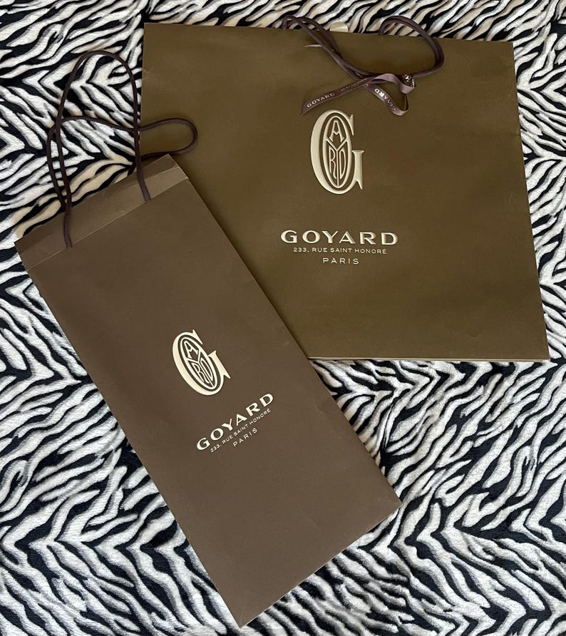 May include: Two brown paper shopping bags with the Goyard logo and address printed in white. The bags have brown handles and are on a black and white zebra-striped background. The text on the bags reads "GOYARD 233, RUE SAINT HONORE PARIS."