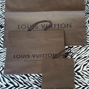 May include: Three brown paper shopping bags with the "LOUIS VUITTON" logo printed in black. The largest bag has a twisted brown handle. The smaller bags also have the brand name and "MAISON FONDEE EN 1854 PARIS" printed on them.