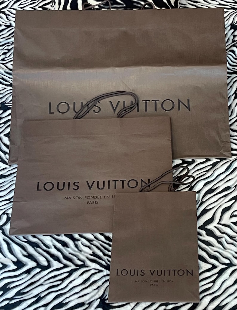 May include: Three brown paper shopping bags with the "LOUIS VUITTON" logo printed in black. The largest bag has a twisted brown handle. The smaller bags also have the brand name and "MAISON FONDEE EN 1854 PARIS" printed on them.