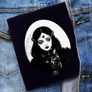 Peut inclure: Illustration en noir et blanc d'une femme aux longs cheveux noirs et d'un chat noir. La femme a un troisième œil et porte une tenue sombre. L'image est sur un fond bleu foncé.