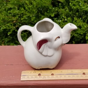 May include: White ceramic creamer in the shape of an elephant with a red painted ear. The creamer has a spout and a handle.