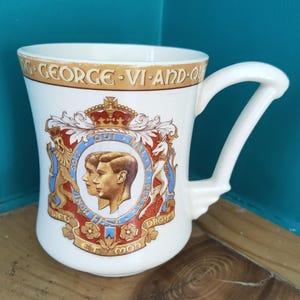May include: A white ceramic mug featuring a royal crest with portraits and the text "GEORGE VI AND QUEEN". The mug has a unique handle design and is decorated with gold, red, blue, and brown accents.