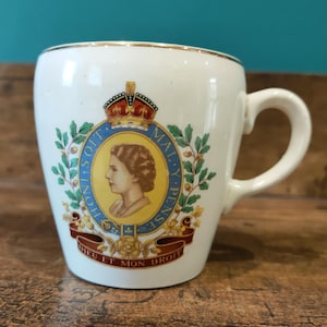 Puede incluir: Taza de cerámica blanca con borde y asa dorados. La taza presenta un emblema real con un retrato, corona y el lema "DIEU ET MON DROIT". El diseño incluye detalles en rojo, azul y dorado.