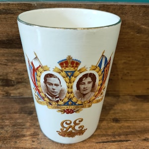 Może przedstawiać: Biały ceramiczny kubek z złotym obramowaniem, upamiętniający koronację z 1937 roku. Zawiera portrety, koronę, flagi i inicjały "GE". Tekst brzmi "Coronation 1937". Zabytkowy przedmiot kolekcjonerski.