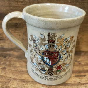 Op de afbeelding: Een beige keramische mok met een gebogen handvat. De mok heeft een gedetailleerd wapen met een kroon, leeuwen en een schild in rood, blauw en goud. Tekst op de mok herdenkt het huwelijk van de Prins van Wales en Lady Diana Spencer, juli 1981.