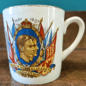 May include: A white ceramic mug with a gold rim, featuring a portrait of George VI and Elizabeth, with the text "MAY 1937" and "GEORGE VI ELIZABETH CORONATION". The mug has a curved handle and is decorated with flags and a royal crest.