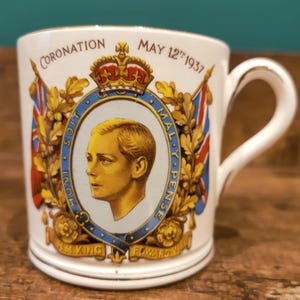 Puede incluir: Taza de cerámica blanca que conmemora la coronación de Eduardo VIII el 12 de mayo de 1937. Presenta un retrato de Eduardo VIII, banderas y las palabras "Coronation" y "May 12th 1937". La taza tiene un borde y un asa dorados.