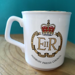 Puede incluir: Taza de cerámica blanca con asa, con un emblema dorado y rojo de una corona y el texto "The Queen's Silver Jubilee 1952-1977 Modbury Parish Council". La taza tiene un borde dorado y un asa blanca.