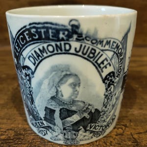 May include: A white and blue commemorative cup with a portrait of Queen Victoria. The cup is inscribed with "Leicester Diamond Jubilee Commemoration Queen Victoria 1897". The cup is cylindrical in shape.