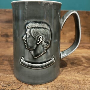 May include: Gray ceramic mug with a handle, featuring a profile of a person and the text "H.R.H. THE PRINCE OF WALES" on a banner. The mug has a glossy finish and a white rim.