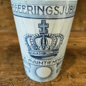 May include: A white and blue ceramic cup with a crown and cross design. The cup has the words "REGERINGSJUBIL" and "JE MAINTIENDRAI" printed on it. The design is in a classic blue and white color scheme.