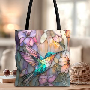 May include: Tote bag featuring a colorful hummingbird design with a stained-glass effect. The bag showcases a vibrant hummingbird with blue, green, and orange hues, surrounded by purple and pink flowers. Black straps complete the design.