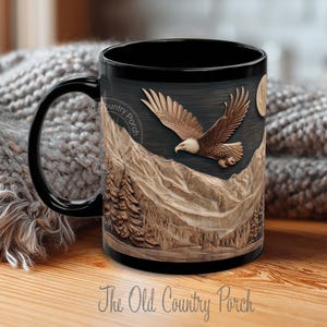 May include: Black ceramic mug featuring a detailed relief carving of an eagle in flight over a mountain range, with a full moon in the background. The mug has a black handle and the text "The Old Country Porch".