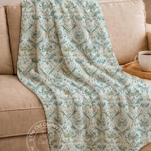 May include: A floral patterned throw blanket in shades of blue, green, and cream is draped over a beige sofa. The blanket features a repeating damask design. The Old Country Porch logo is visible in the lower left corner.