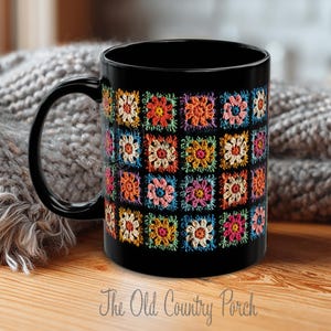 May include: Black ceramic mug featuring a colorful crochet floral pattern. The design includes squares with flowers in shades of orange, pink, yellow, and blue. The mug has a black handle and the text "The Old Country Porch" at the bottom.