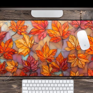 May include: A rectangular mouse pad featuring a stained-glass style design of autumn maple leaves in shades of orange, red, and yellow. A white computer mouse rests on the pad. The pad is on a wooden desk with a keyboard and monitor.
