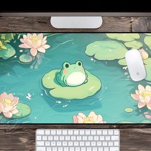 May include: A rectangular mousepad featuring a cartoon illustration of a green frog sitting on a lily pad in a pond. Pink lotus flowers and lily pads surround the frog. A white mouse and keyboard are also visible.