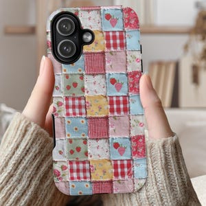 May include: A patchwork phone case featuring a mix of floral, gingham, and strawberry patterns in red, pink, blue, and yellow. The case has a black border and a camera cutout. The case is held by a person wearing a beige sweater.